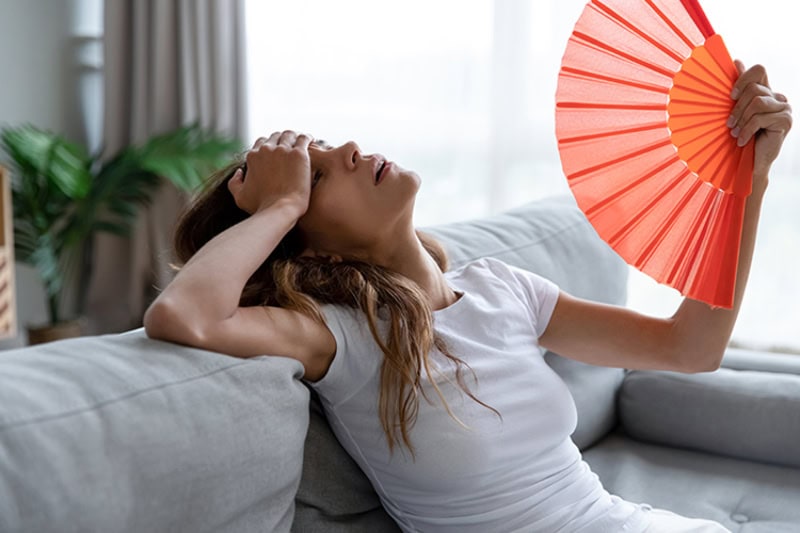 side view of an exhausted young girl using a paper fan to cool down in her apartment.