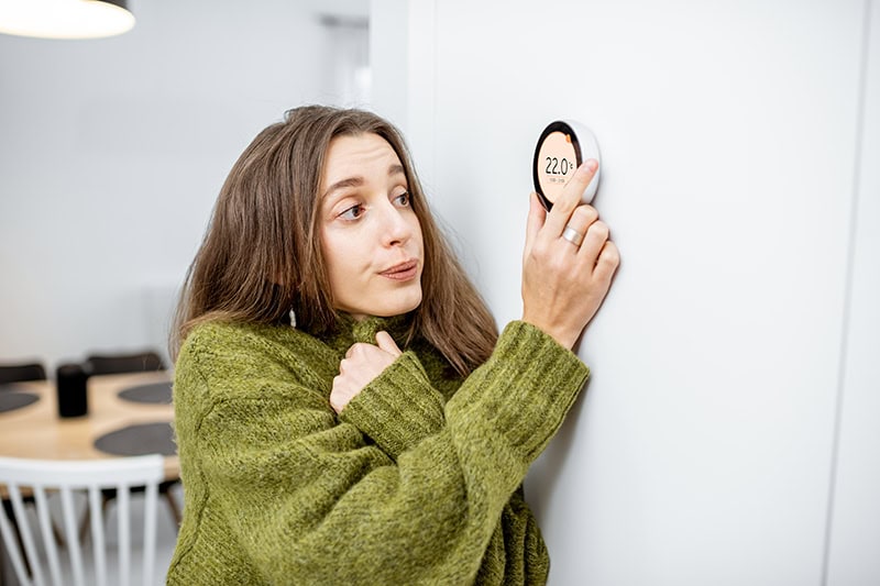 woman shivering in her sweater as she turns up the thermostat.